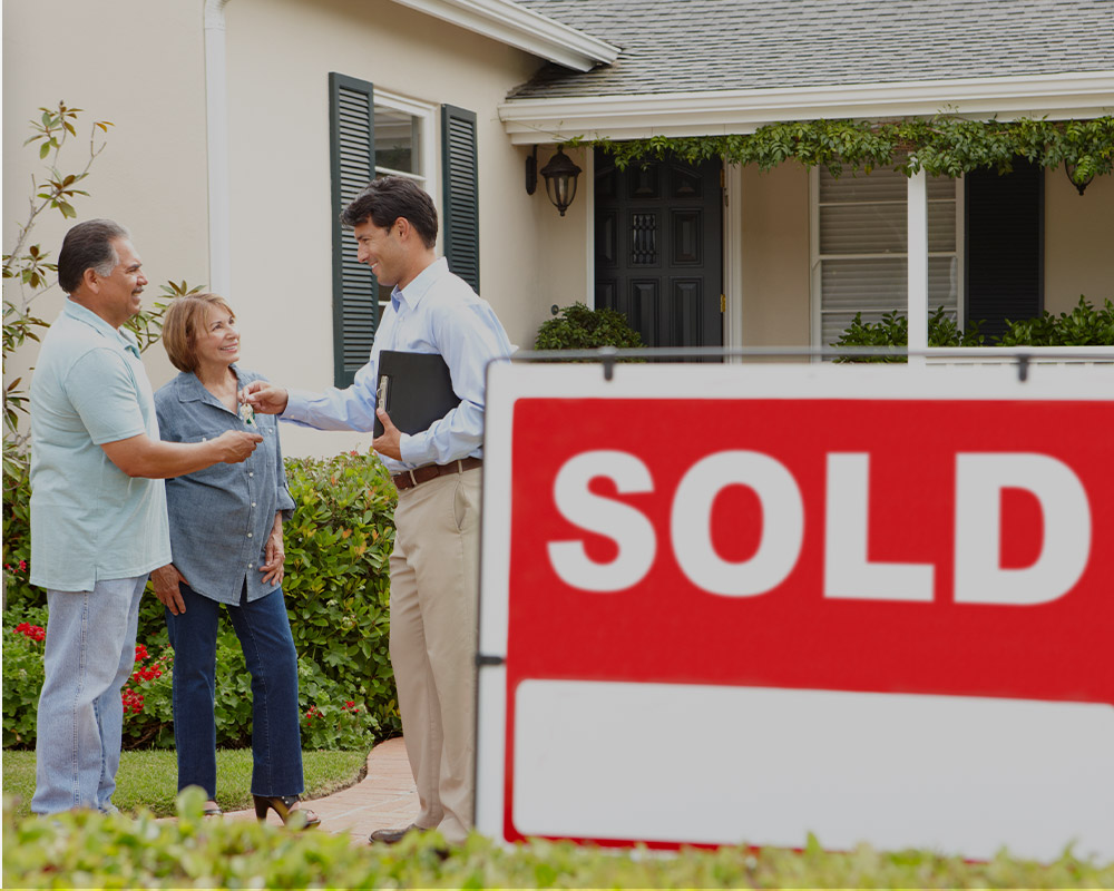 Realtor handing house keys to a couple with a “Sold” sign in front.