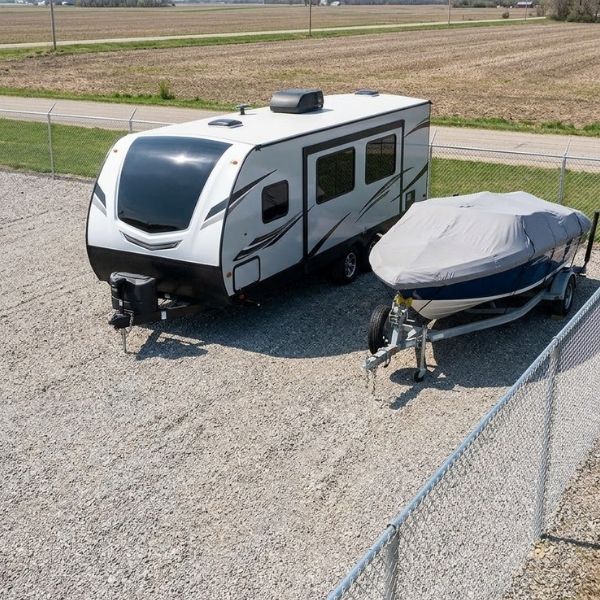 A gravel storage lot holds a camper trailer and a covered boat within a secure fence.